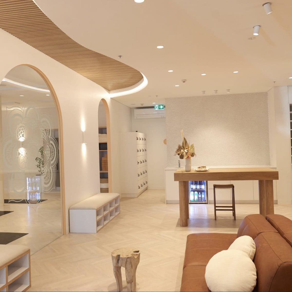 A modern interior space featuring a beige couch, wooden furniture, greenery, and light-colored walls with curved architectural details.