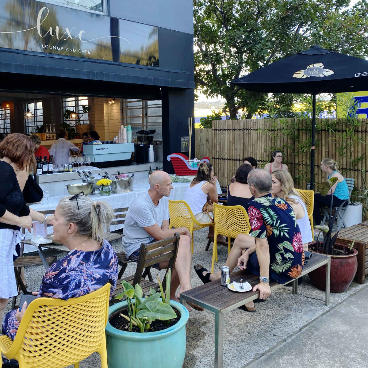 People are seated at outdoor tables in a cafe, socializing and enjoying food and drinks in a cozy courtyard setting.