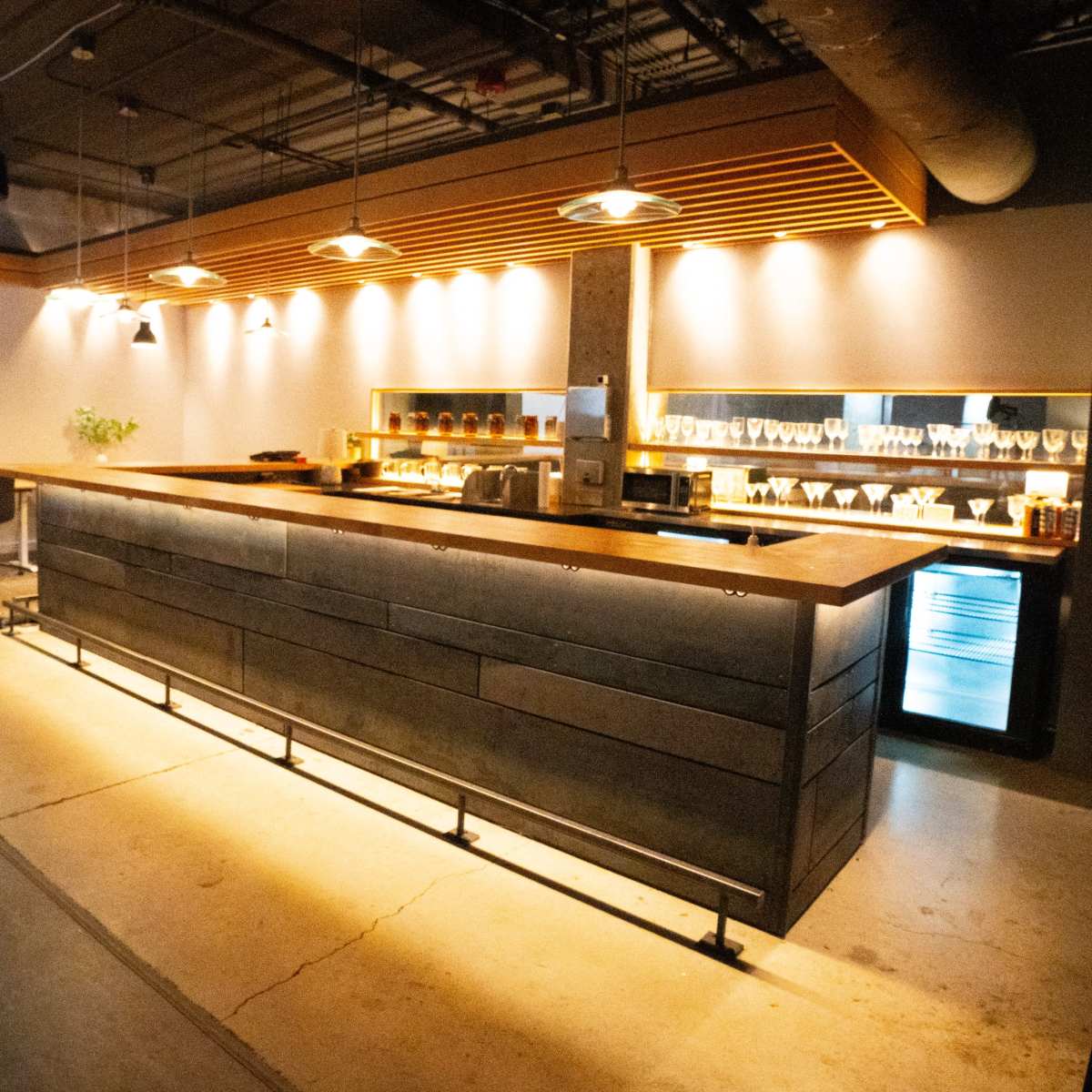 A modern bar area with a wooden countertop, illuminated by overhead lights, and shelves displaying glassware and a refrigerator.