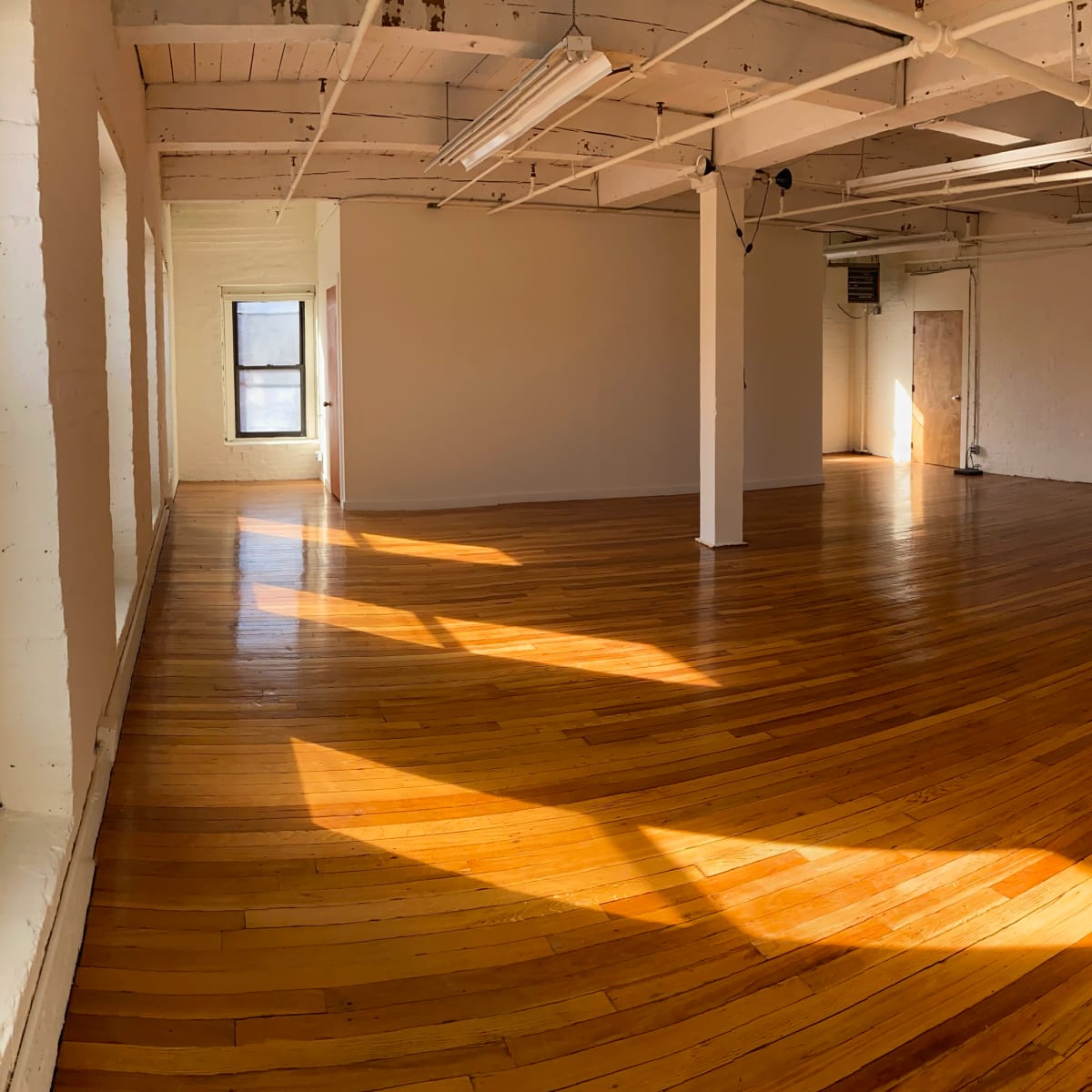 A spacious, empty room with wooden floors and large windows allowing sunlight to illuminate the space.