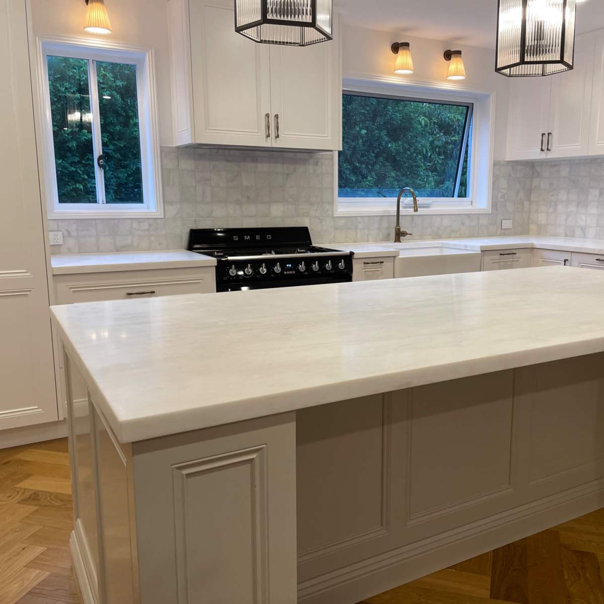 A modern kitchen featuring a large white marble island, stainless steel appliances, and light-colored cabinetry with a herringbone wood floor.