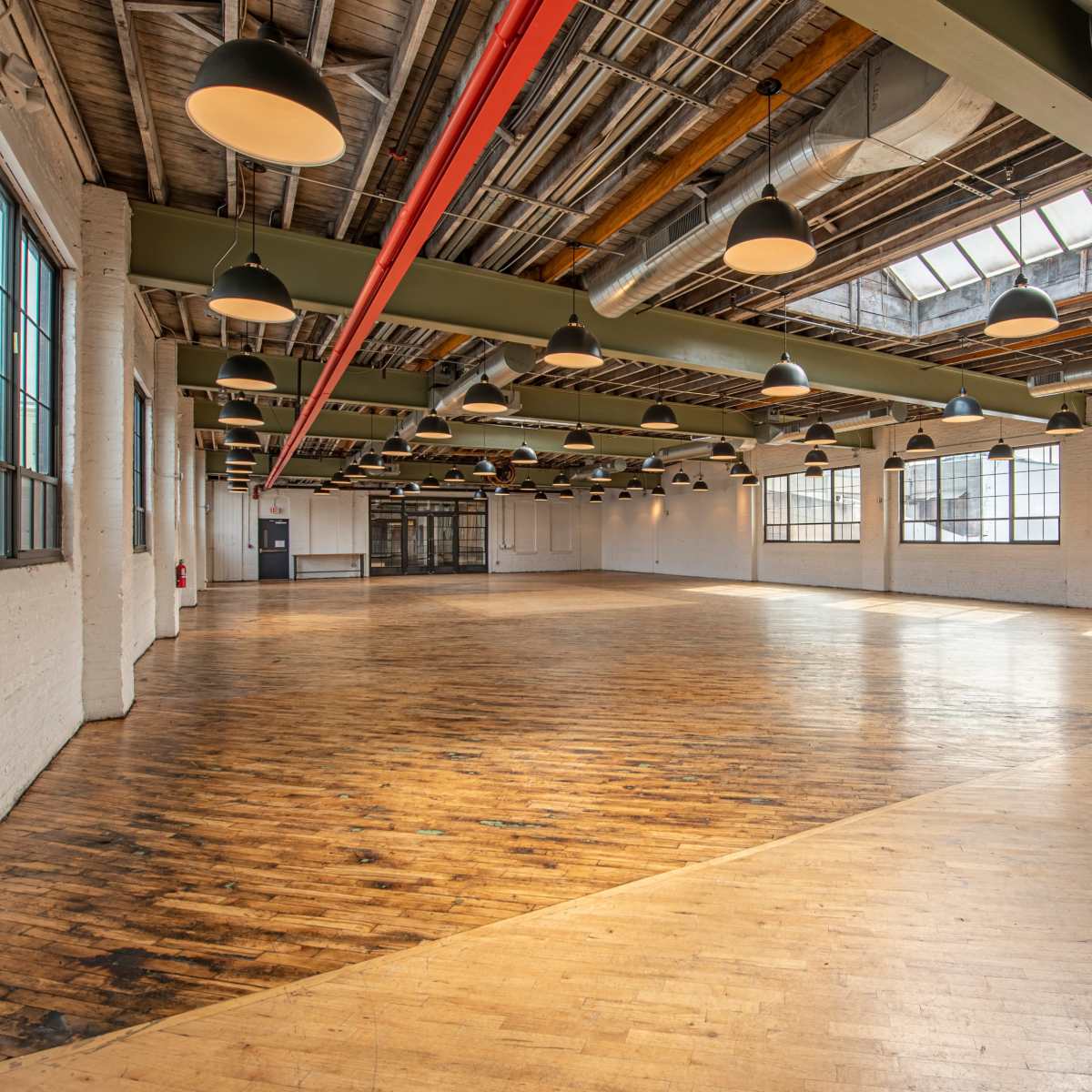 A spacious, empty industrial-style room with wooden floors, large windows, and hanging light fixtures.