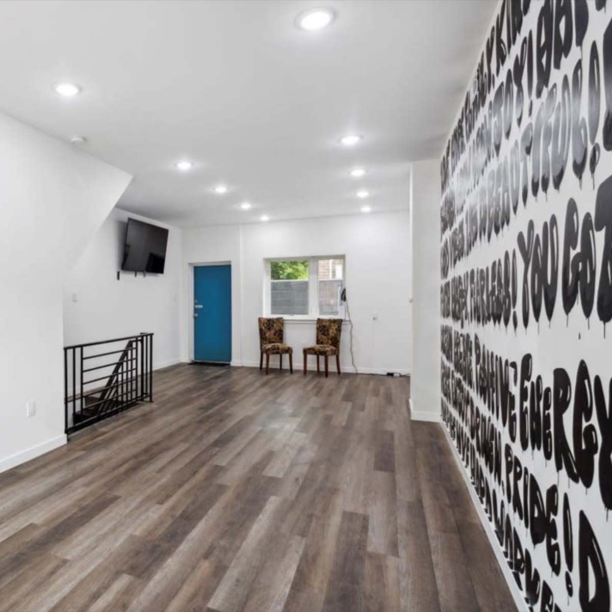 A spacious interior room with hardwood flooring, a wall featuring bold text, two chairs positioned against one wall, and a blue door at the far end.