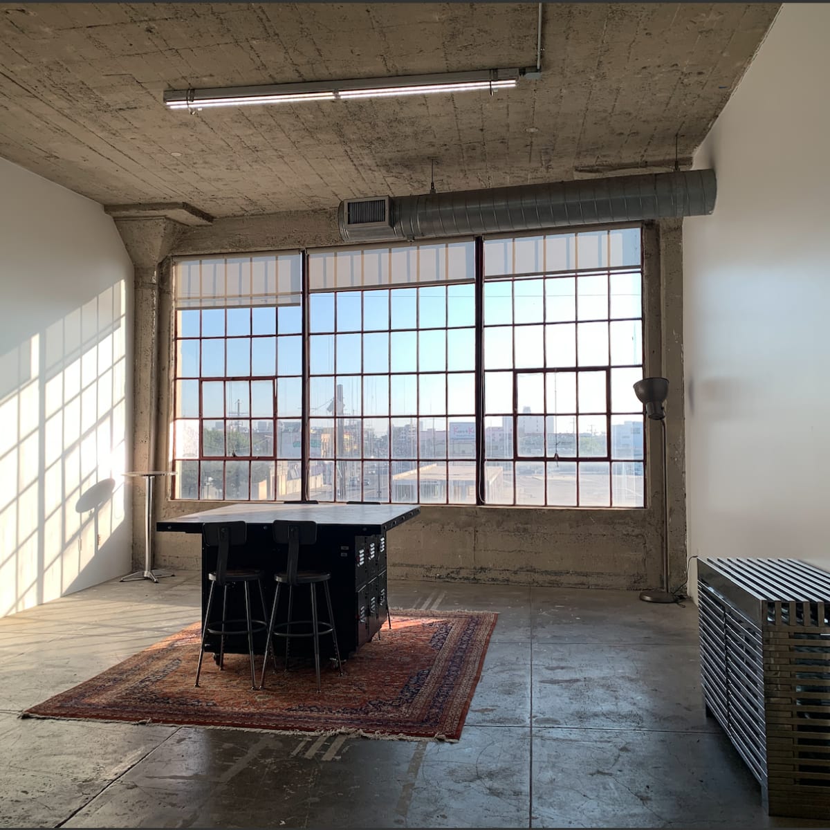 A spacious room with large windows, concrete walls, a rug, a table with stools, and an arcade machine in the corner.