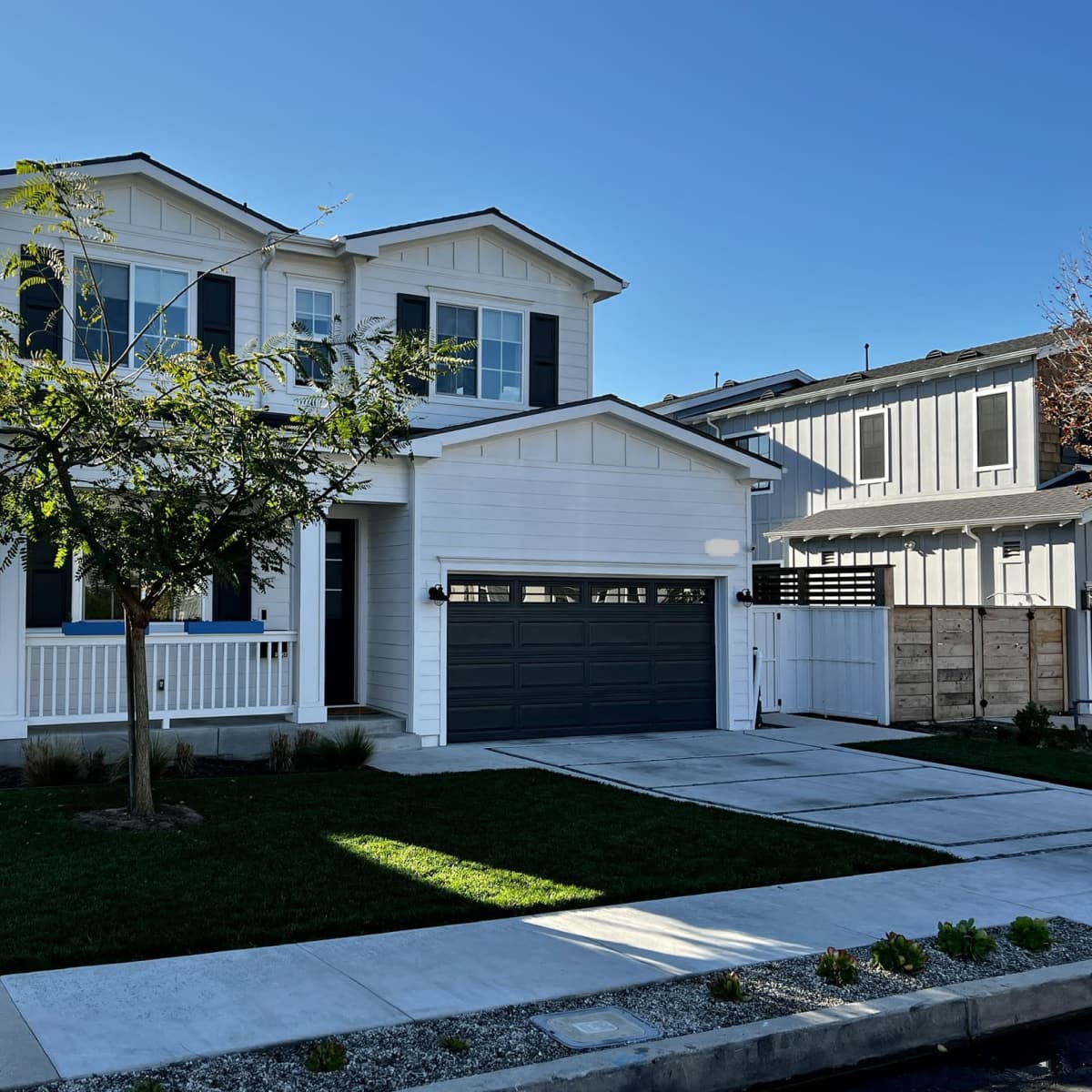 A two-story white house with black shutters and a gray garage door is situated on a landscaped corner lot.