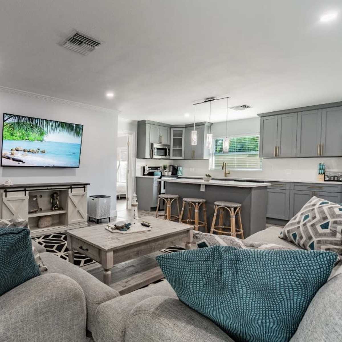 A modern living room with a large TV mounted on the wall, a gray sectional sofa with decorative pillows, a coffee table, and an open kitchen area in the background.