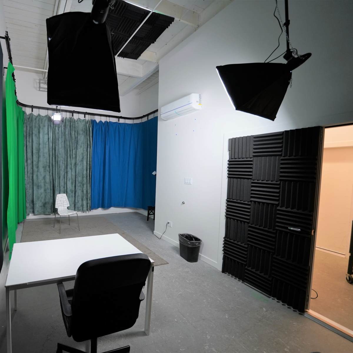 A blank studio room features a white table and chair, green and blue backdrops, and lighting equipment hanging from the ceiling.