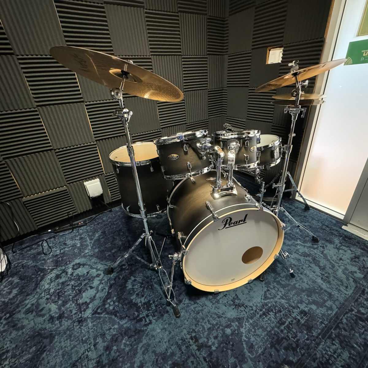 A Pearl drum kit is set up in a soundproofed room with acoustic paneling and a door leading to the outside.