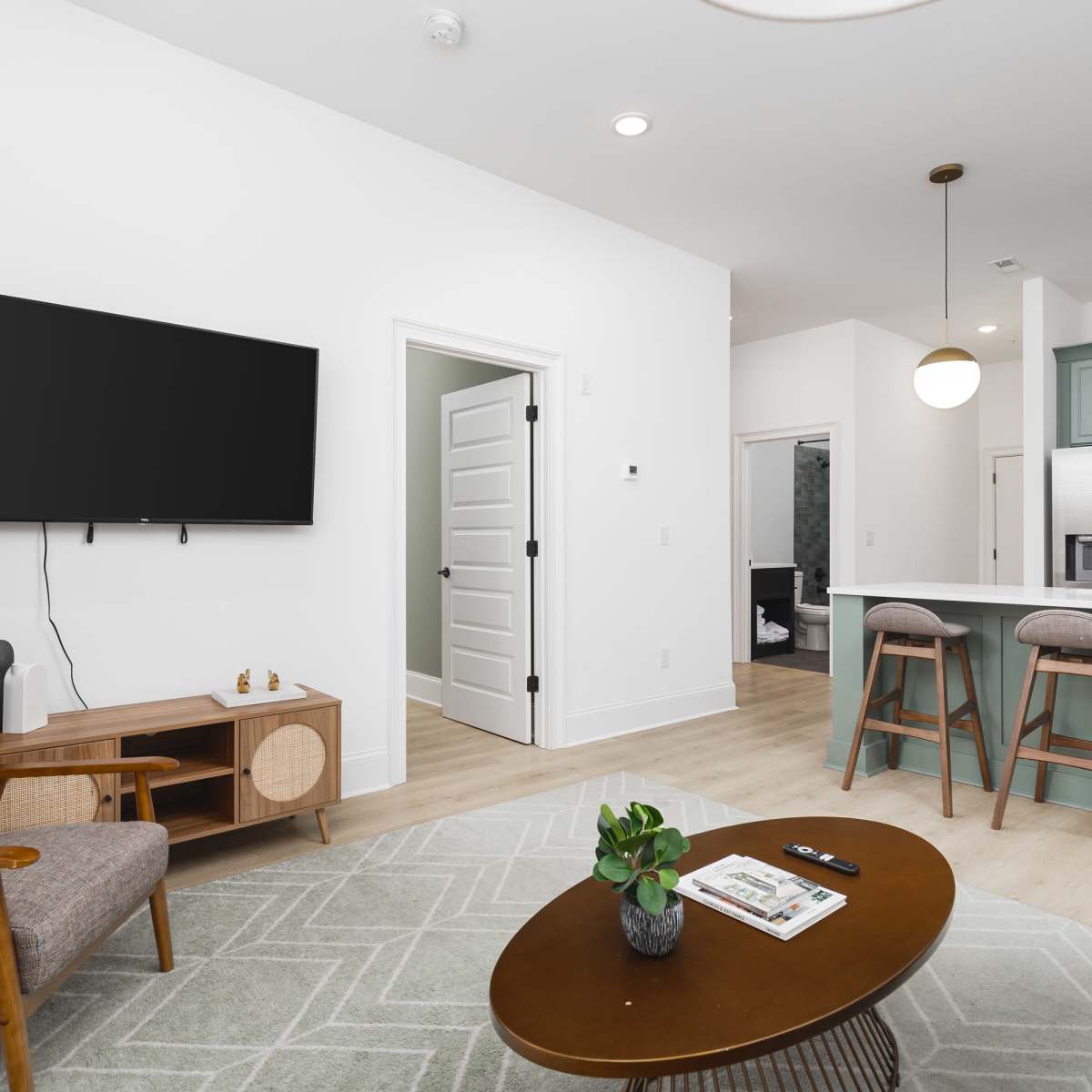 A modern living room with a flat-screen TV mounted on the wall, a seating area with a coffee table and greenery, and a kitchen bar with three stools in the background.