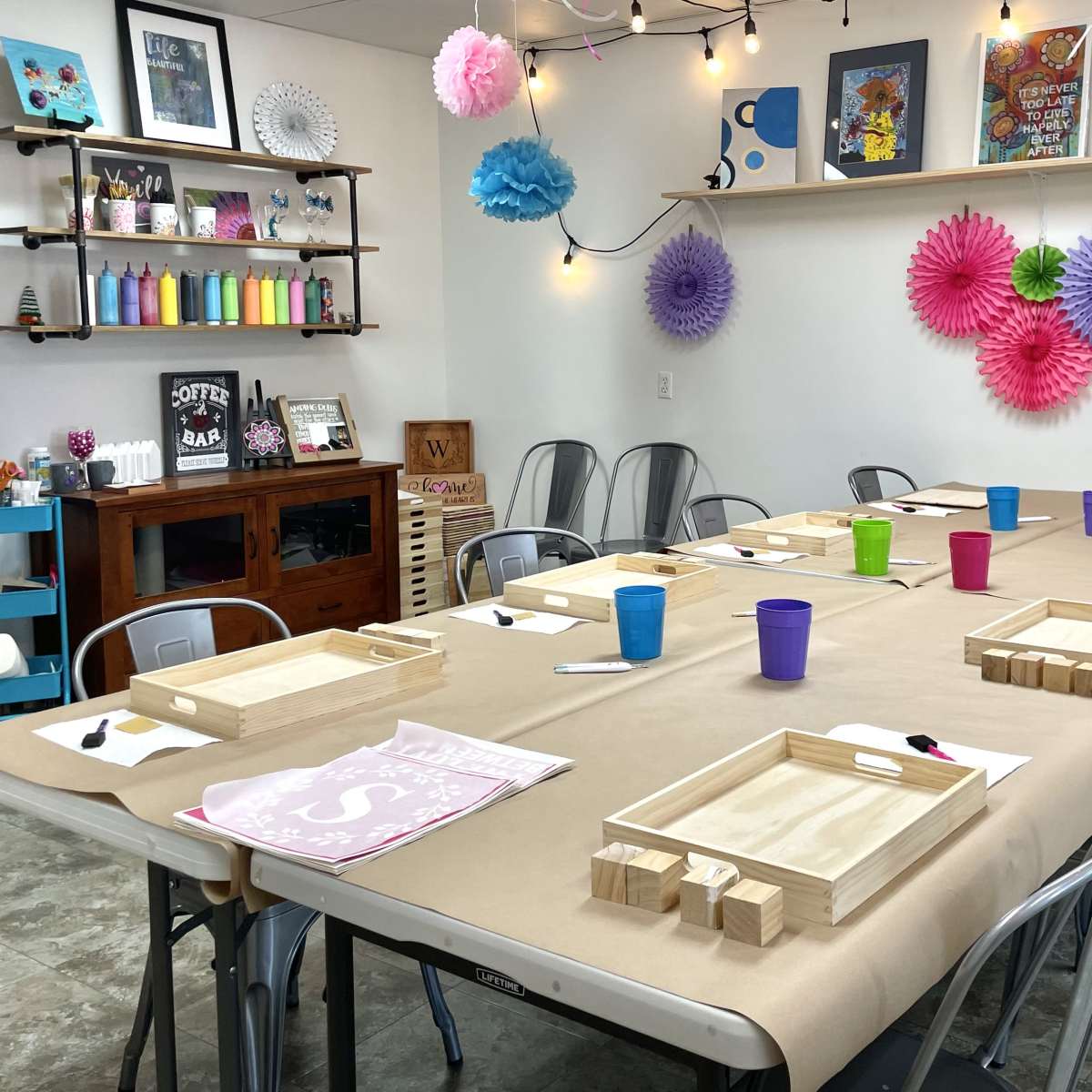 A workshop space with a long table set for an event, featuring colored cups and wooden trays, surrounded by decorative wall art and paper flowers.