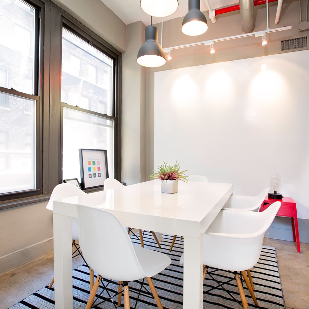A modern meeting room with a white table, four chairs, a potted plant, and a wall with artwork, all illuminated by pendant lighting.