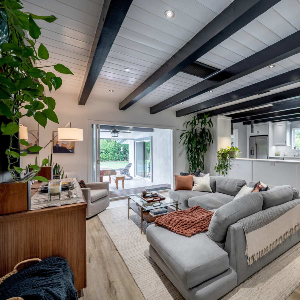 A modern living room features a large sectional sofa, wooden beams on the ceiling, and an open layout leading to a bright kitchen area.