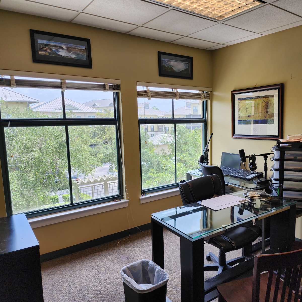An office with a glass-top desk, a chair, and a computer setup near large windows overlooking trees and buildings.
