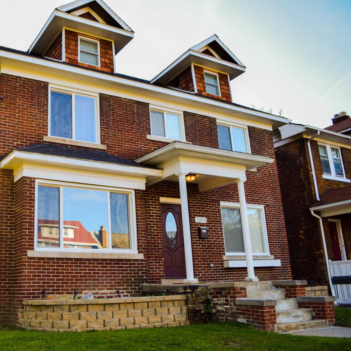 A red brick house with two dormer windows and a large front porch is situated on a grassy lawn.