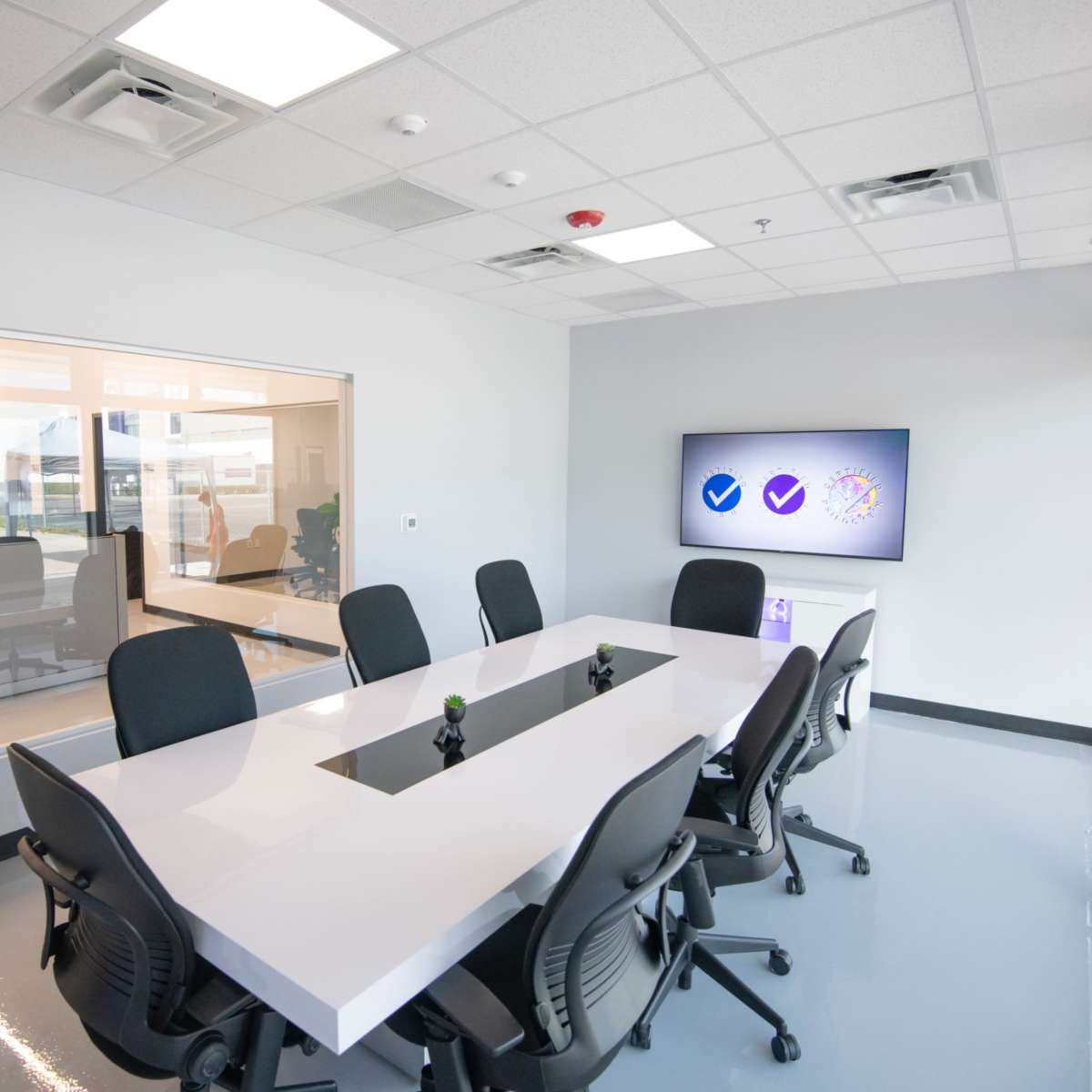 A modern conference room features a large white table surrounded by ergonomic chairs, with a wall-mounted screen displaying logos and a glass wall revealing additional meeting spaces.