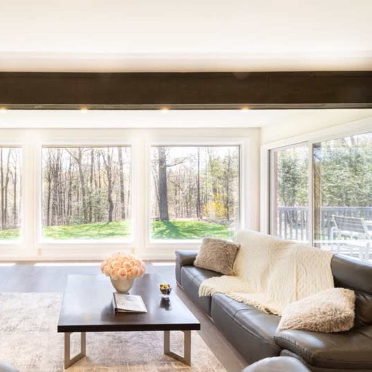 A bright living room features large windows overlooking a wooded area, with a black sofa and a coffee table in the center.