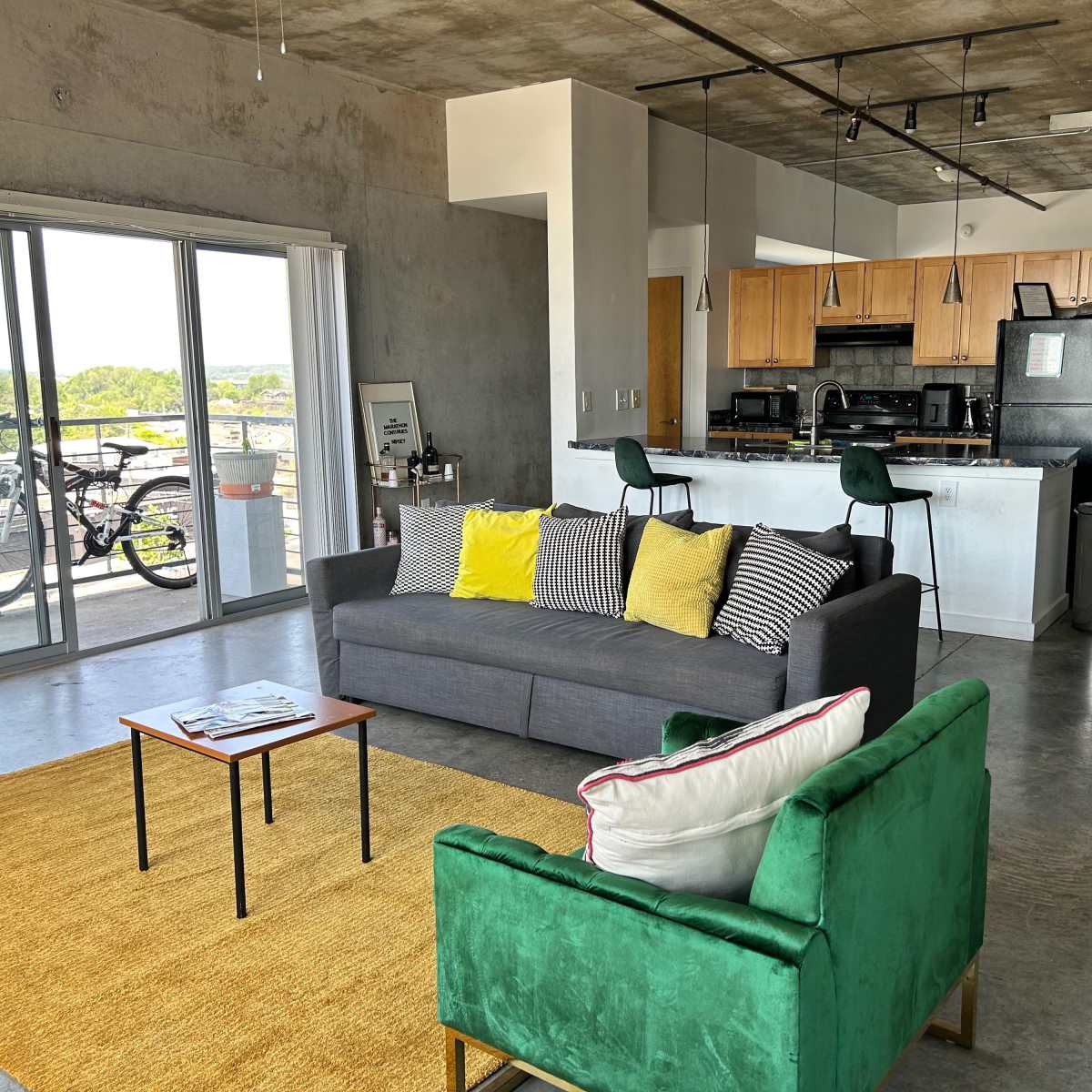 A modern living space with a gray sofa, a green chair, a small coffee table, and a kitchen area, all bathed in natural light from large windows.