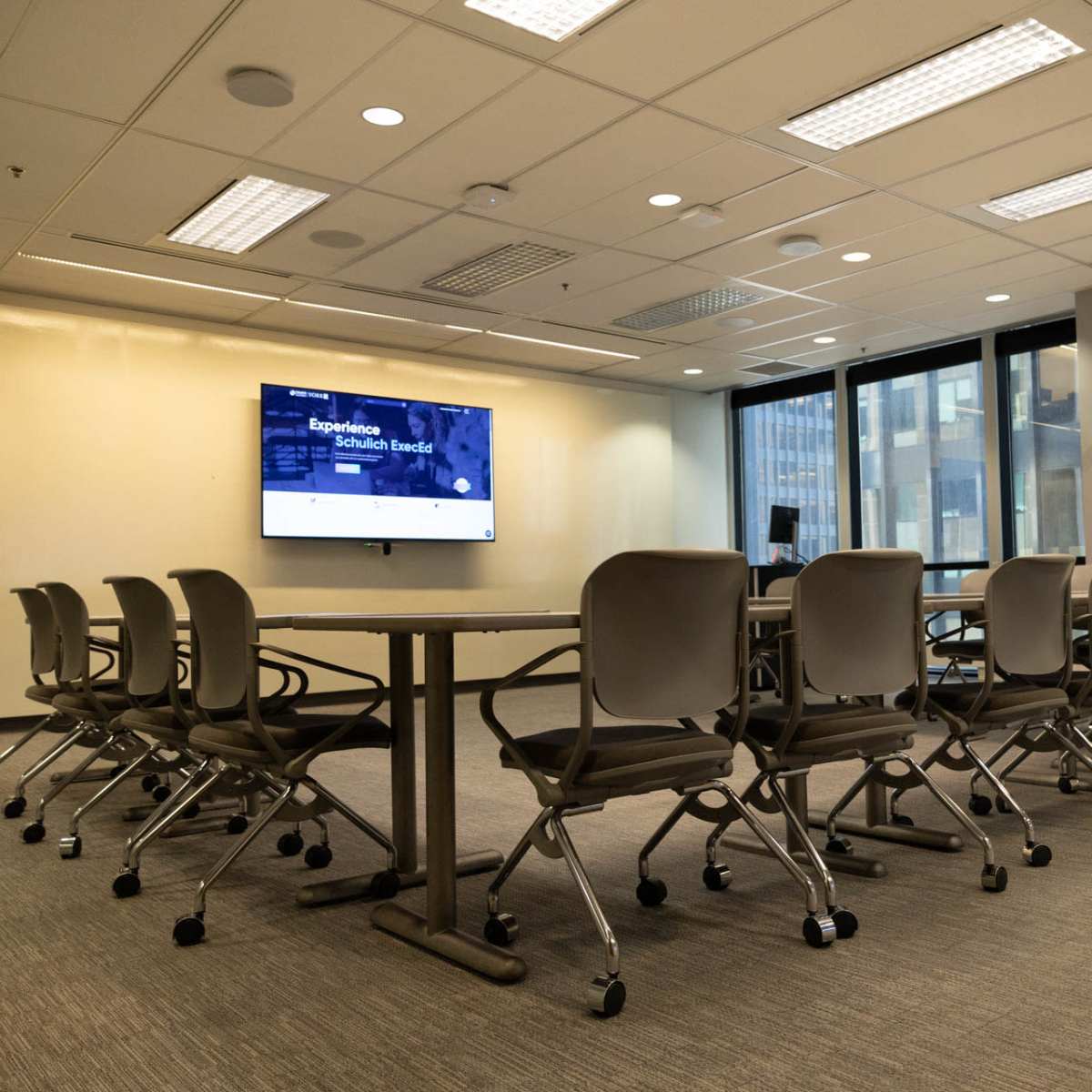 A modern conference room features a large table surrounded by rolling chairs, with a screen displaying a digital presentation on one wall and windows allowing natural light.