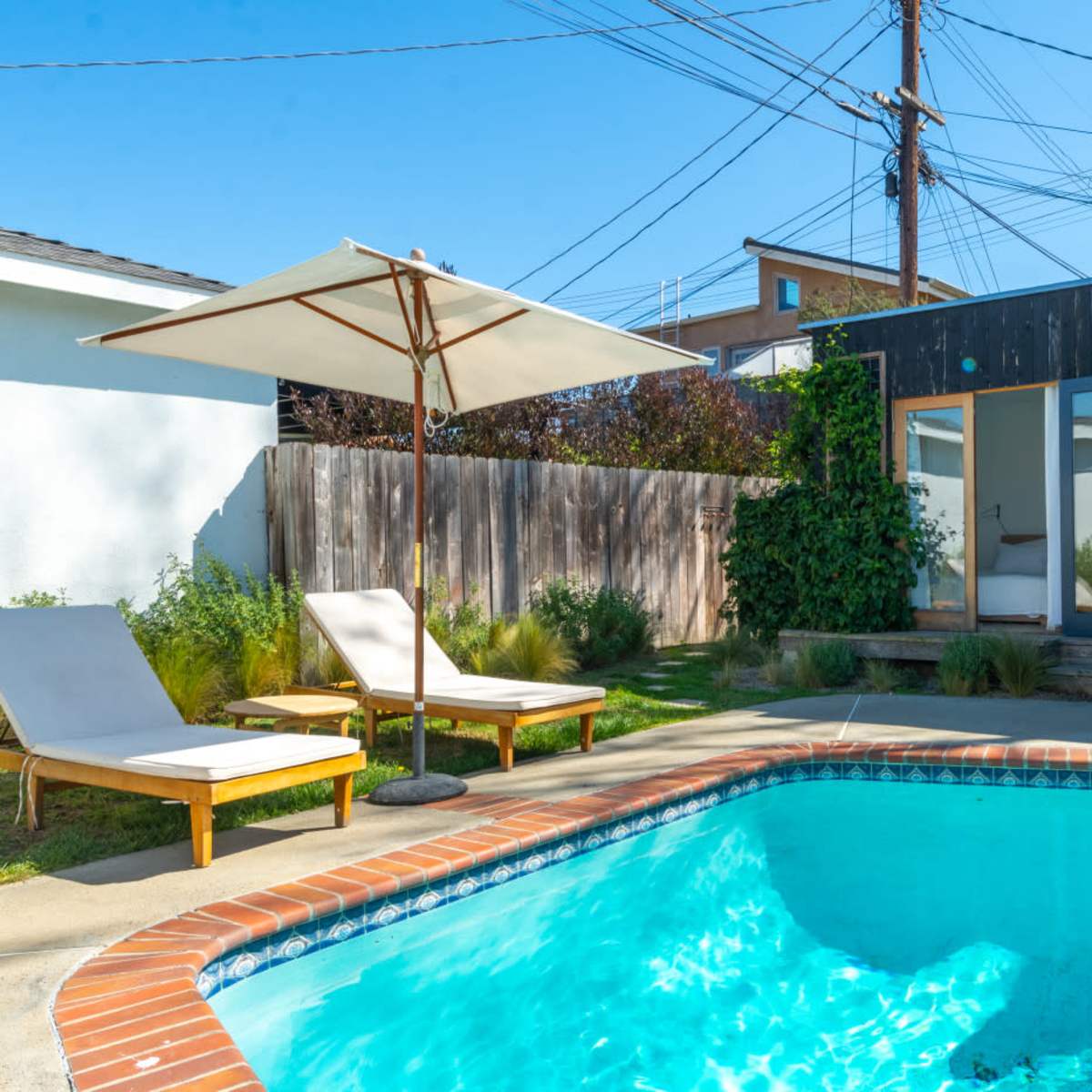 A rectangular swimming pool is surrounded by two lounge chairs and a patio umbrella, with a modern building and greenery in the background.