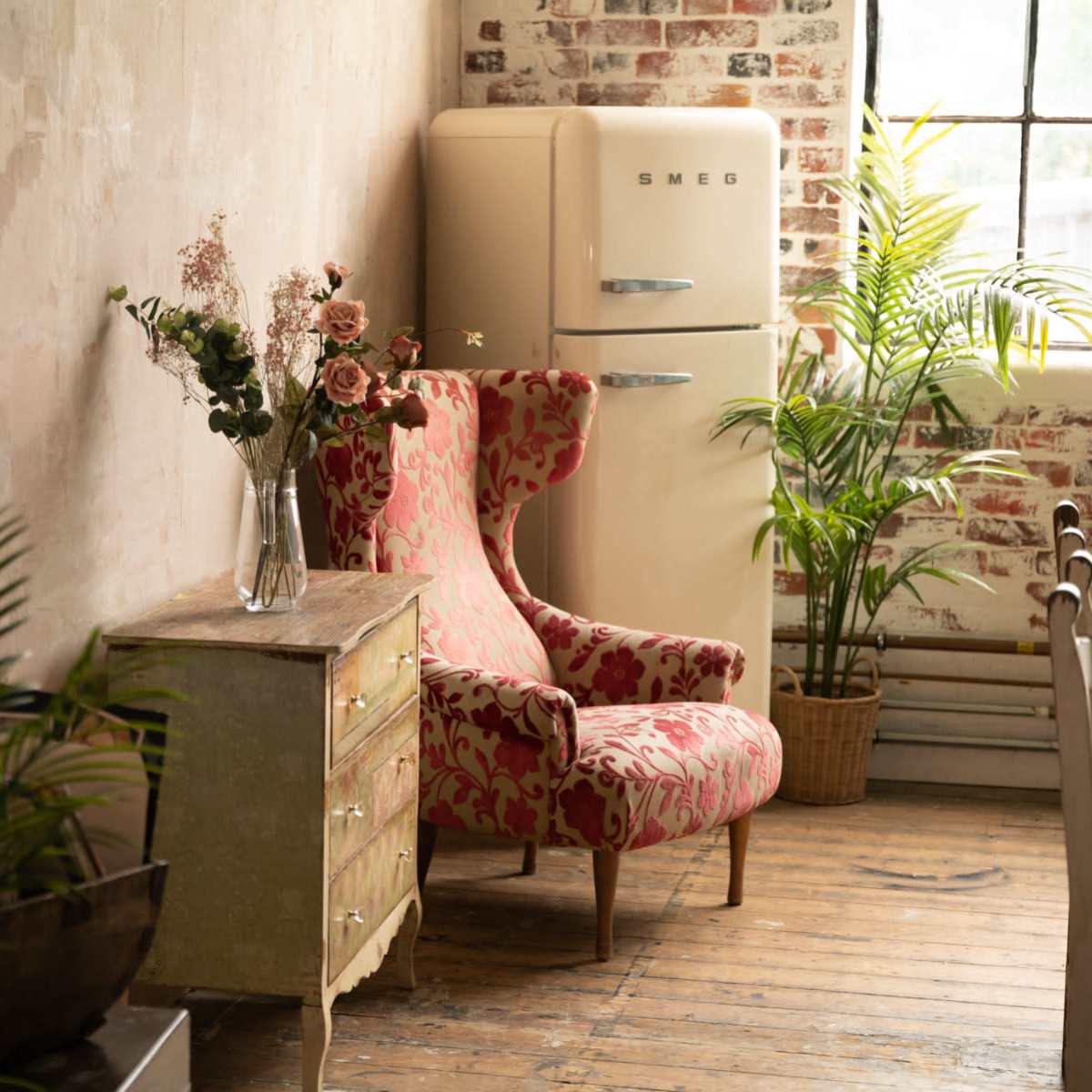 A cozy interior features a vintage Smeg refrigerator, a floral armchair, a wooden chest, and potted plants against a textured wall.