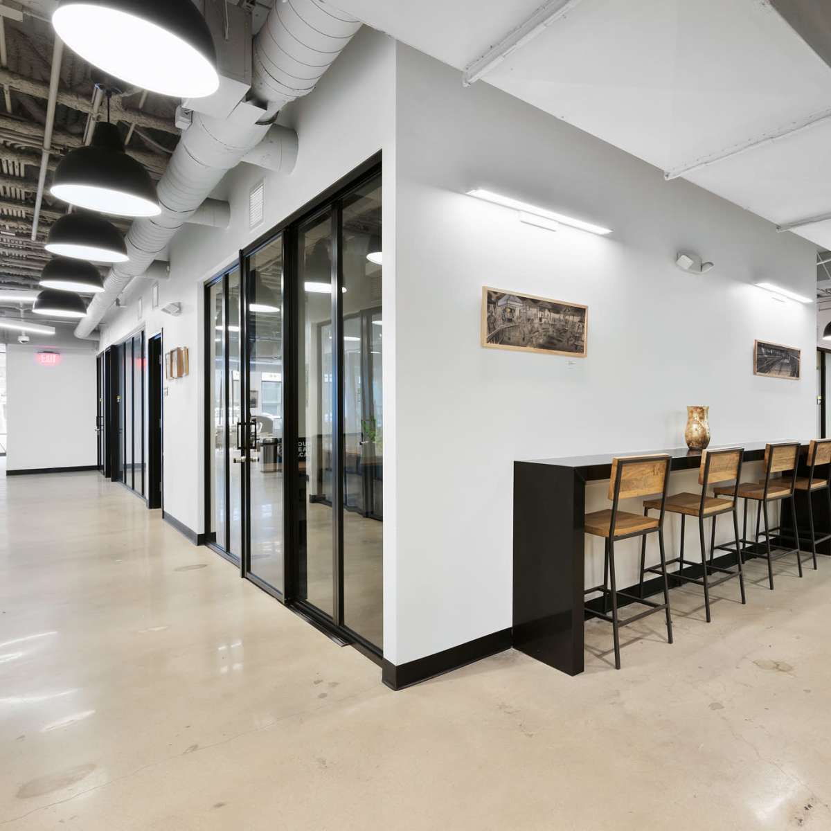 A modern office space featuring a row of high stools at a counter, with glass-walled rooms and doorways leading to other areas.