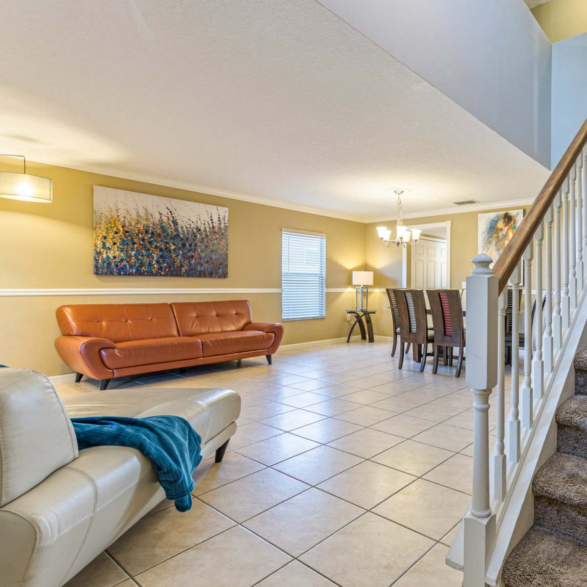 A living area with a staircase leading to an upper level, featuring a couch, a dining table, and artwork on the walls.
