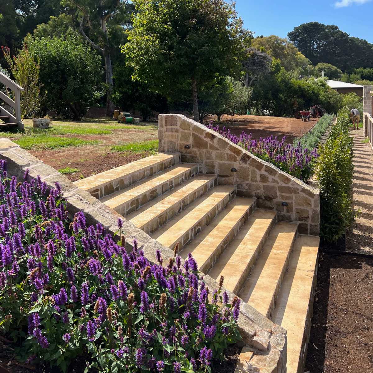 A stone staircase leading to a garden with flowering plants and trees under a clear blue sky.