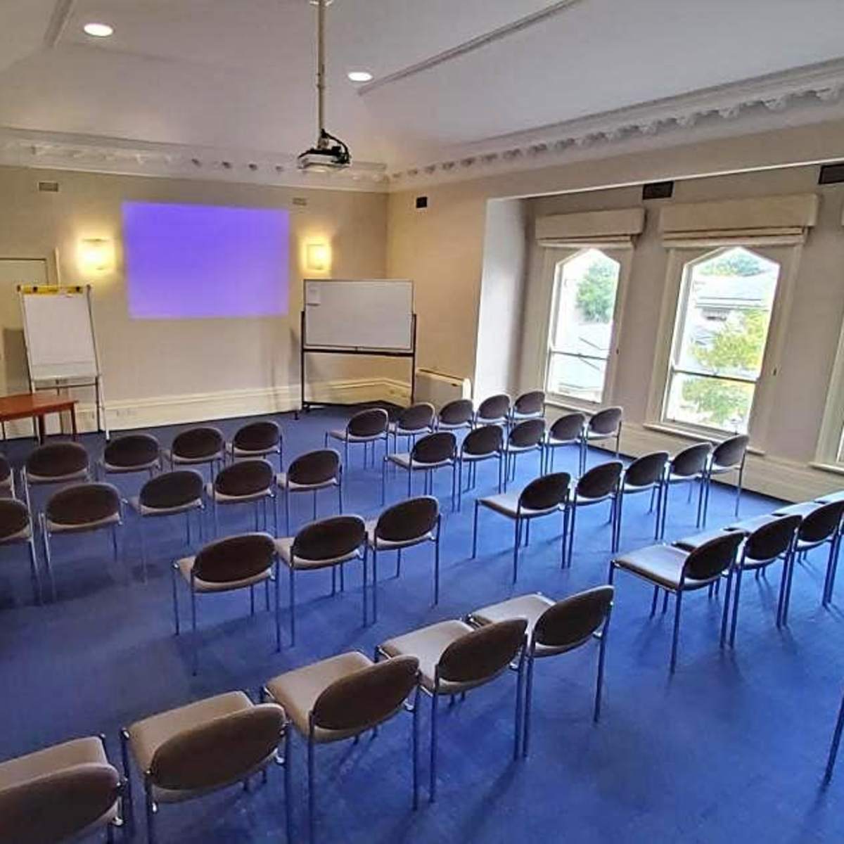 A conference room with rows of chairs facing a blank projection screen, flanked by windows and a table at the back.
