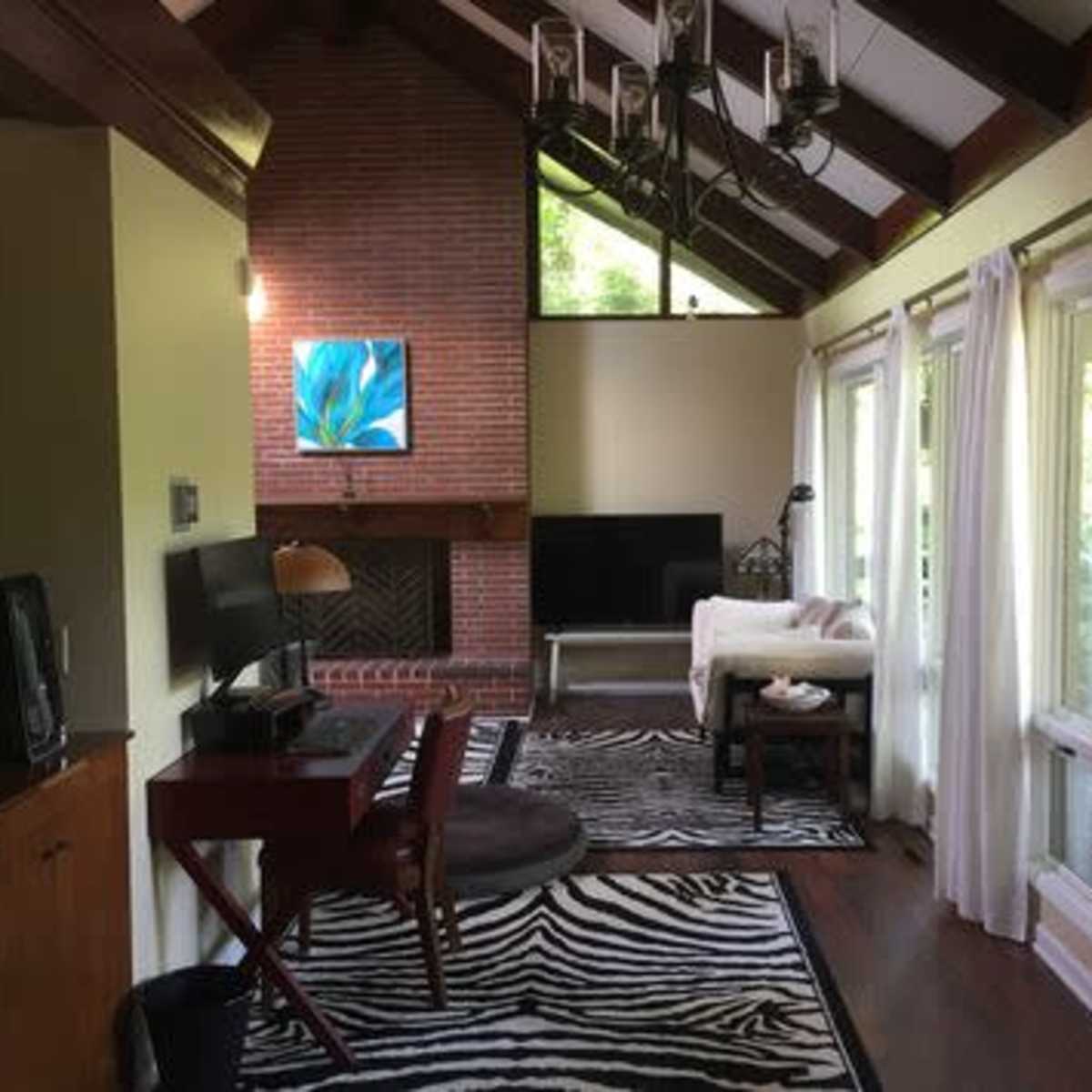 A living room with a high ceiling, featuring a brick fireplace, a mounted artwork, a television, and large windows leading to a wooden deck.