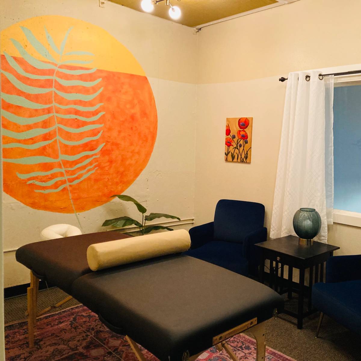 A treatment room featuring two massage tables, a blue armchair, a small side table, and a large orange wall mural of a leaf.