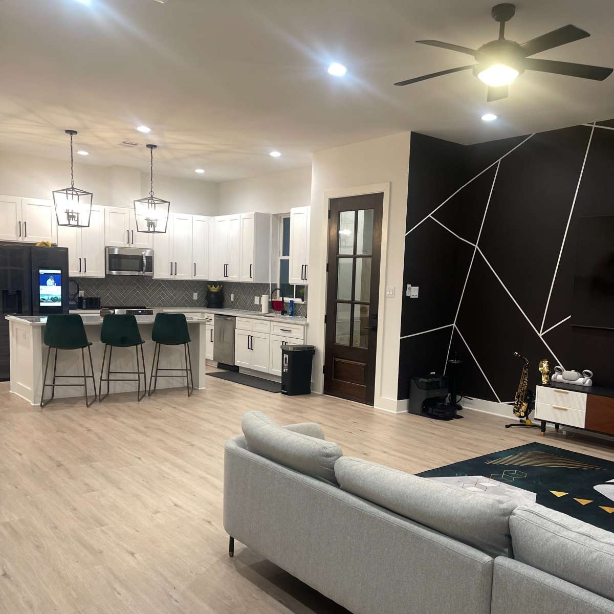 A modern kitchen and living area with white cabinets, bar seating, and a dark geometric accent wall.