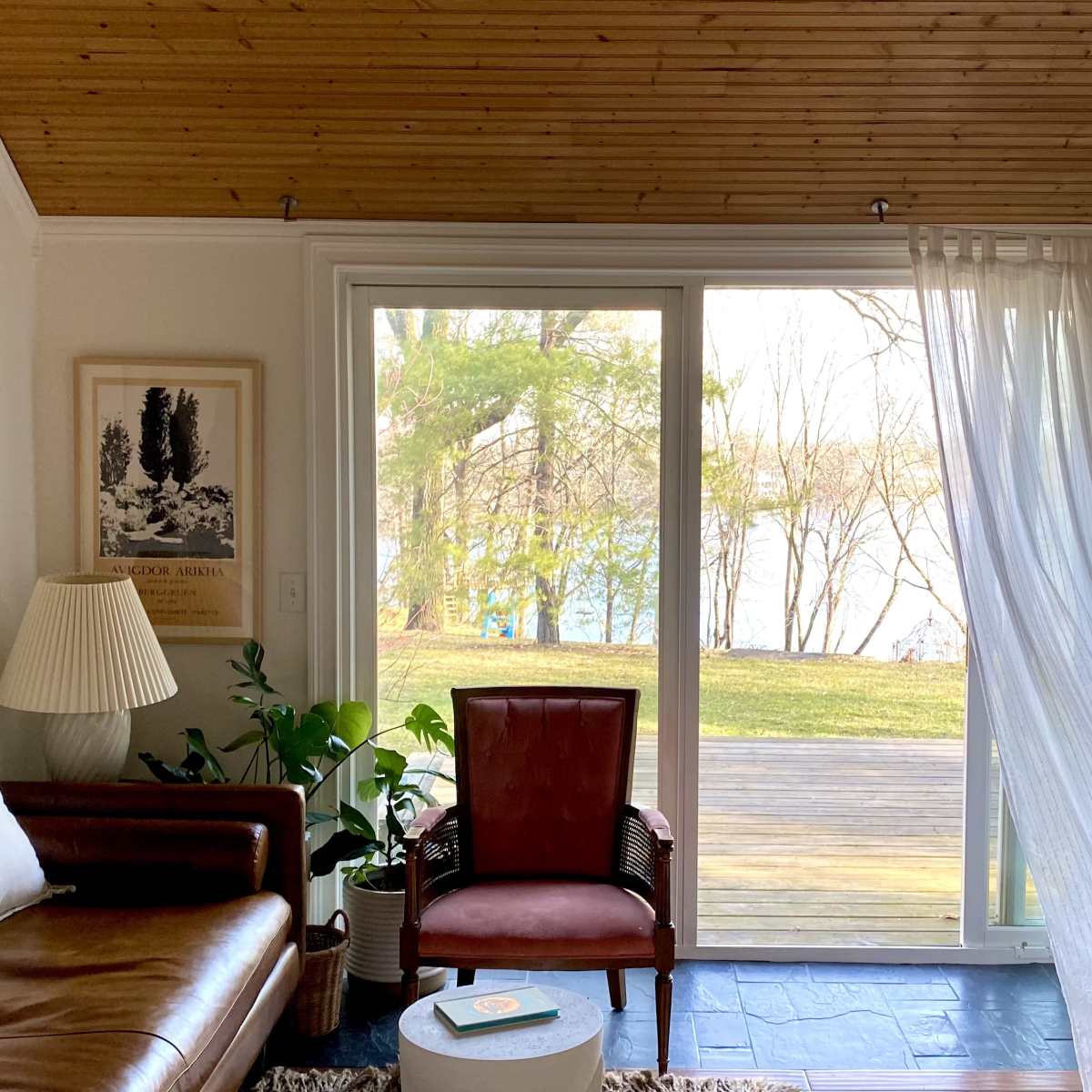 A cozy living room features a brown leather sofa, a red armchair, and large windows overlooking a lake, with a wooden ceiling and light curtains.