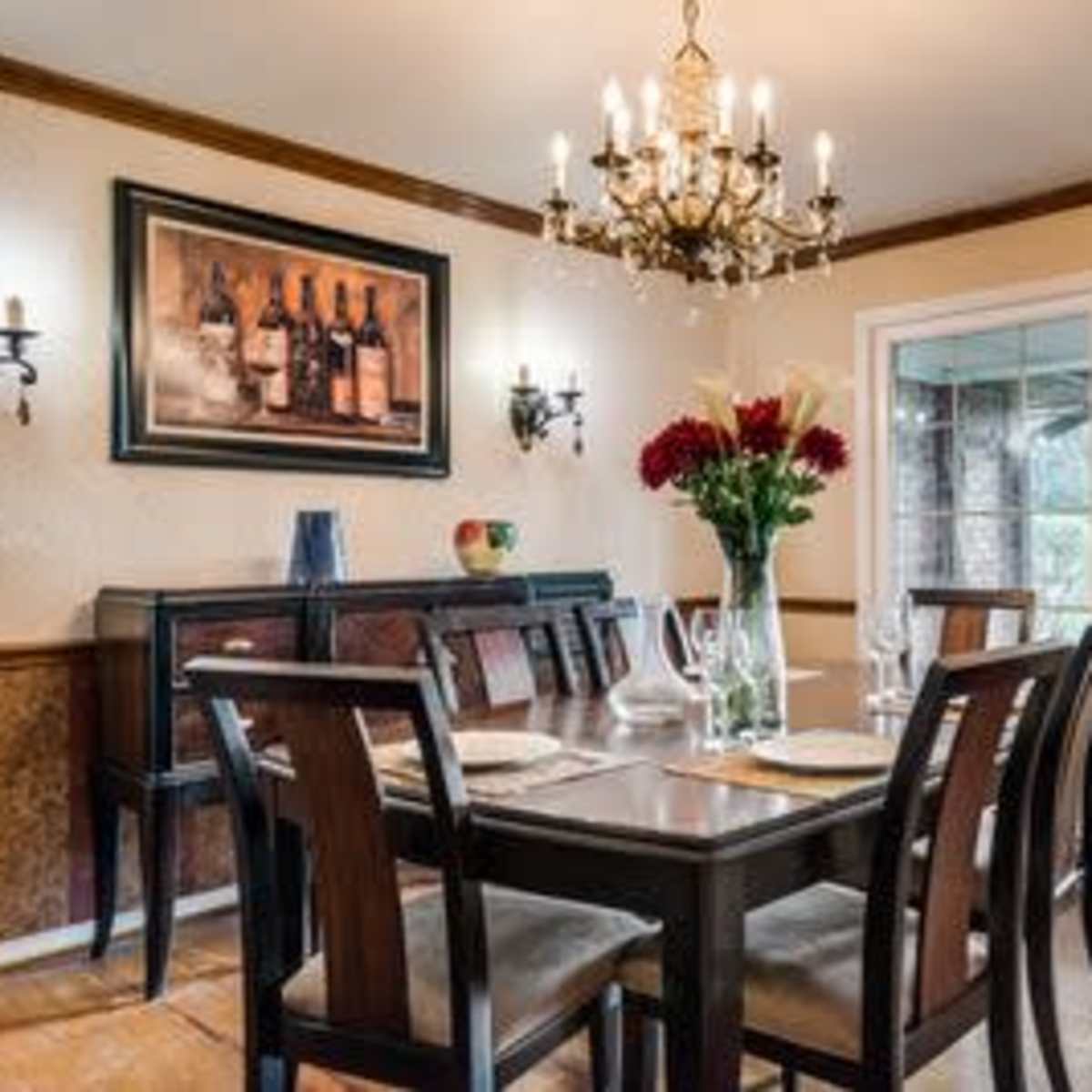 A dining room features a wooden table set for dinner, surrounded by chairs, with a chandelier hanging overhead and a large window looking out to a green landscape.