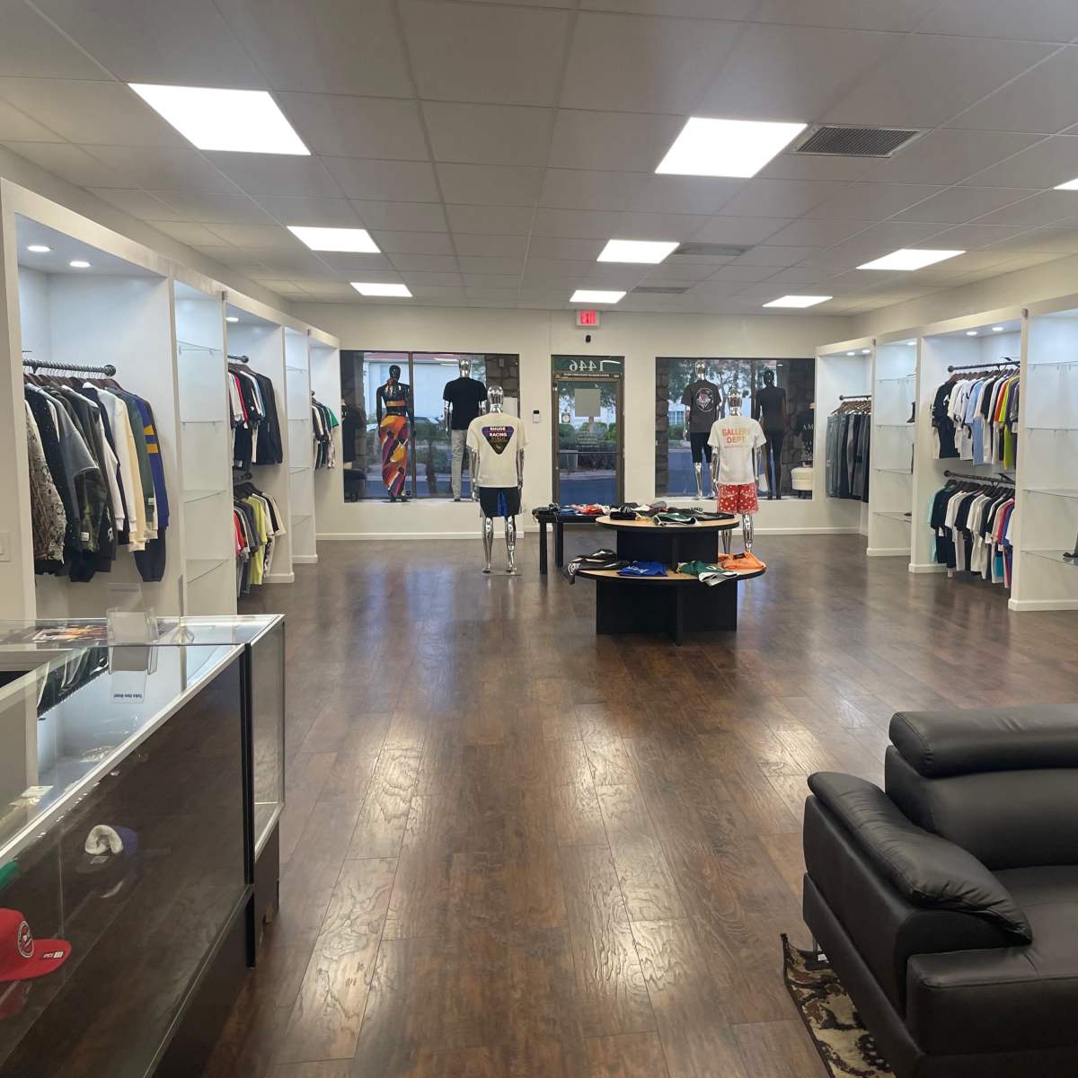A retail clothing store featuring neatly arranged racks of various apparel along the walls, with a central display table and a seating area.