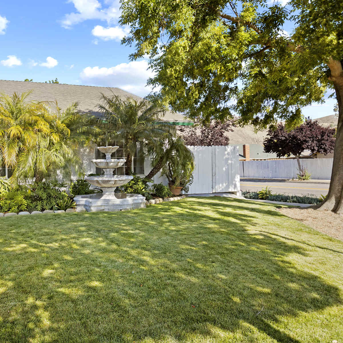 A well-maintained front yard featuring a fountain, lush greenery, and a pathway leading to a single-story house.