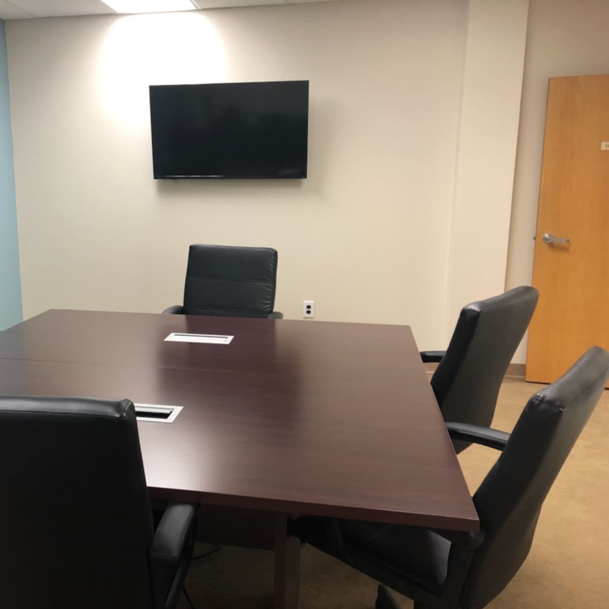 A vacant conference room features a large wooden table surrounded by black leather chairs, with a wall-mounted television and a door in the background.