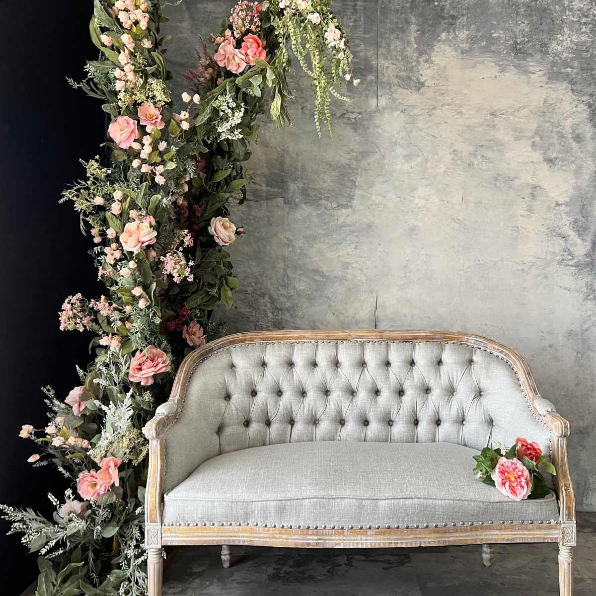 A tufted gray couch is positioned in front of a vertical arrangement of flowers and greenery against a textured gray backdrop.
