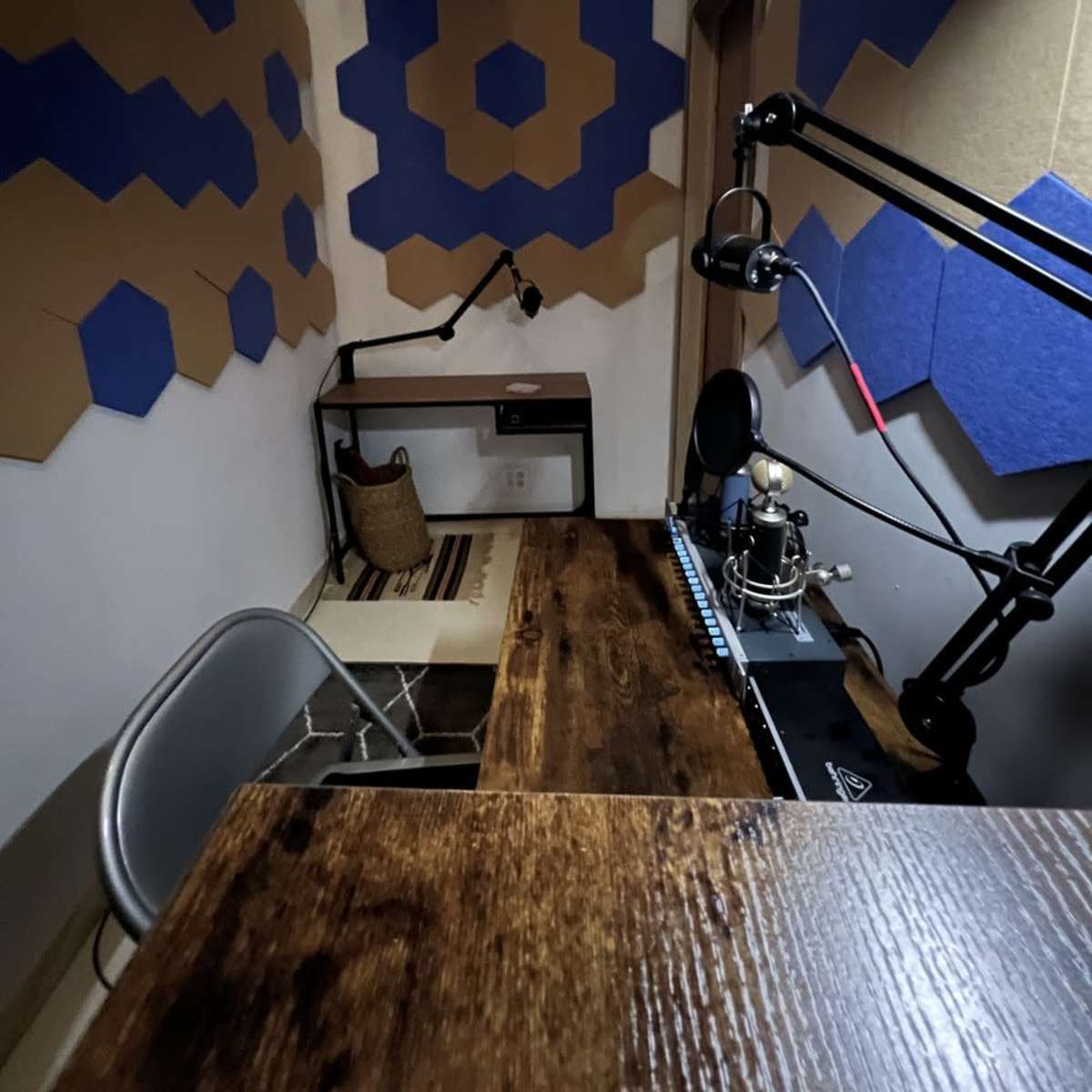 A small recording studio featuring a wooden desk, a microphone setup, and soundproofed walls with blue and tan hexagonal patterns.
