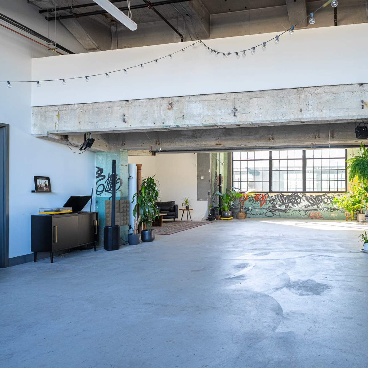 An open, industrial-style interior space with large windows, polished concrete floors, and greenery alongside a graffiti-covered wall.