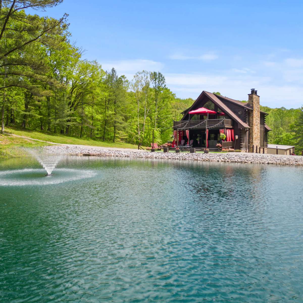 A rustic house with a stone chimney overlooks a pond featuring a fountain, surrounded by lush green trees.