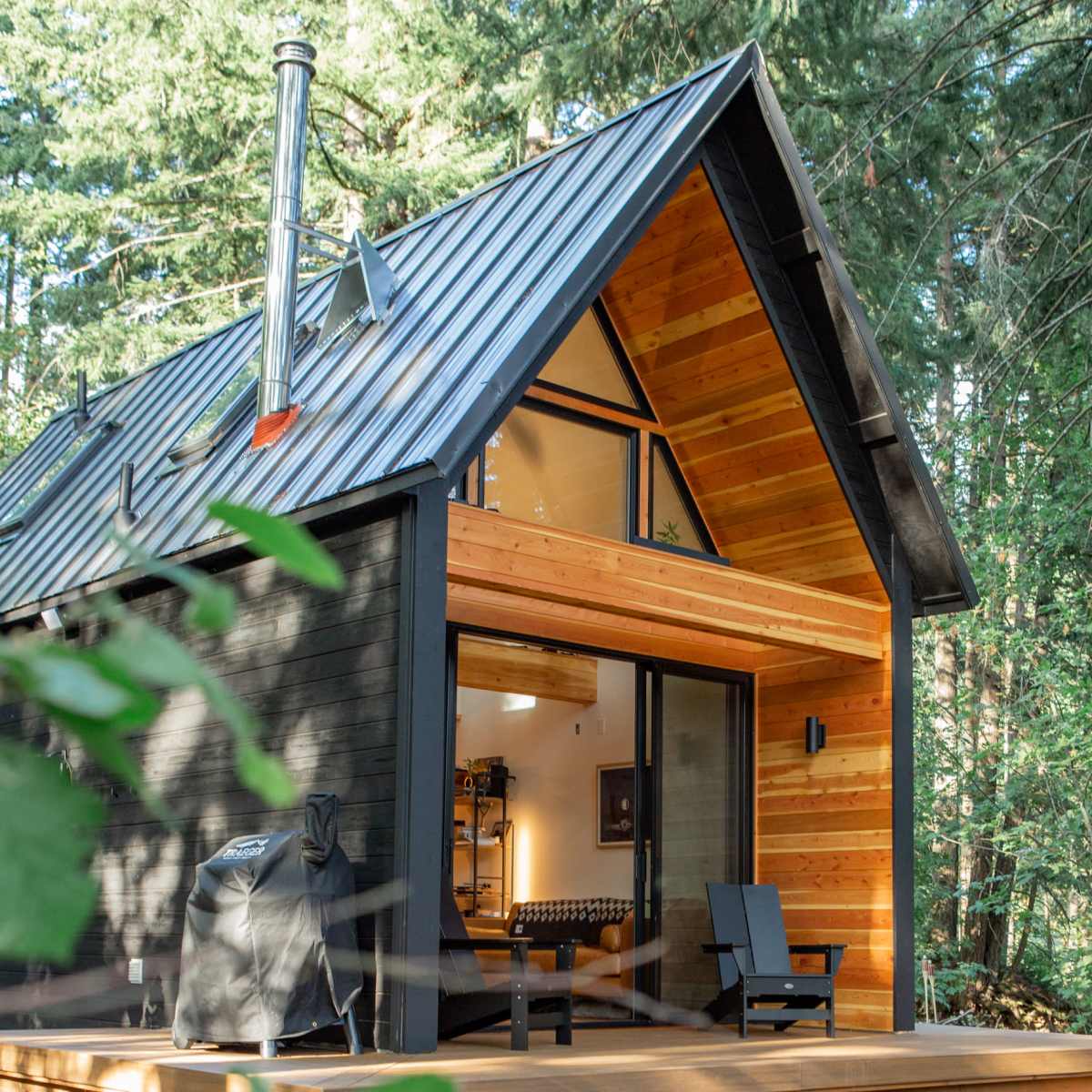 A modern cabin with a sloped metal roof and wooden accents is set amidst tall trees, featuring large glass doors that open onto a wooden deck.