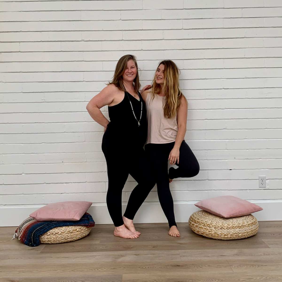 Two women stand next to each other against a white brick wall, with woven poufs and cushions on the floor.
