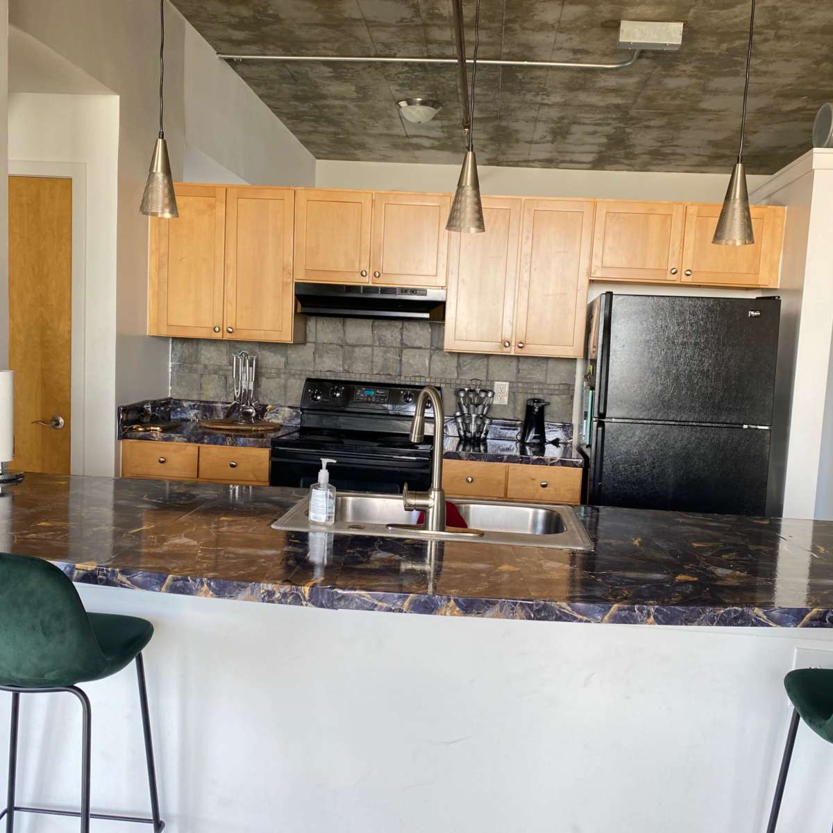 A modern kitchen with wooden cabinets, a black refrigerator, and a marble countertop with two green bar stools.