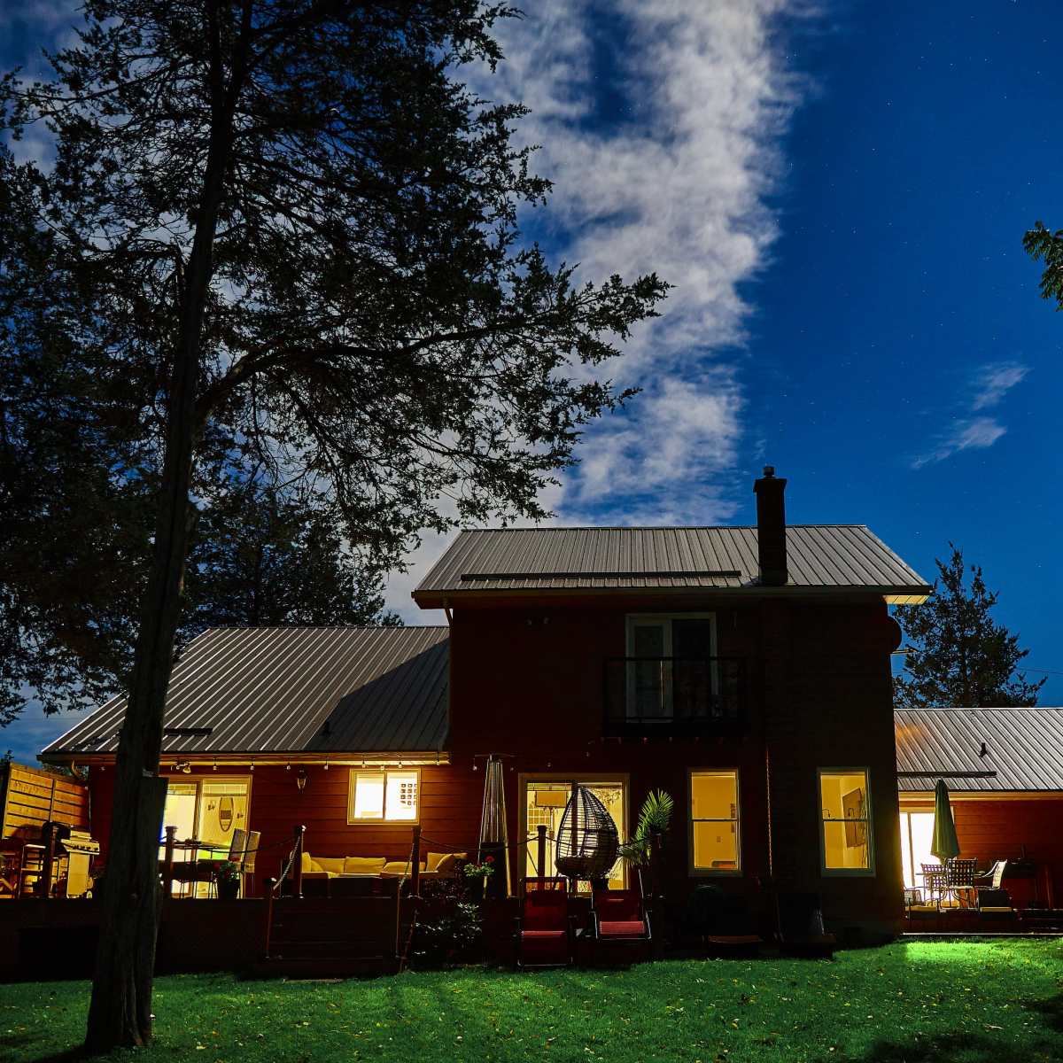 A red house with a metal roof is illuminated at night, surrounded by trees and a clear sky dotted with stars.