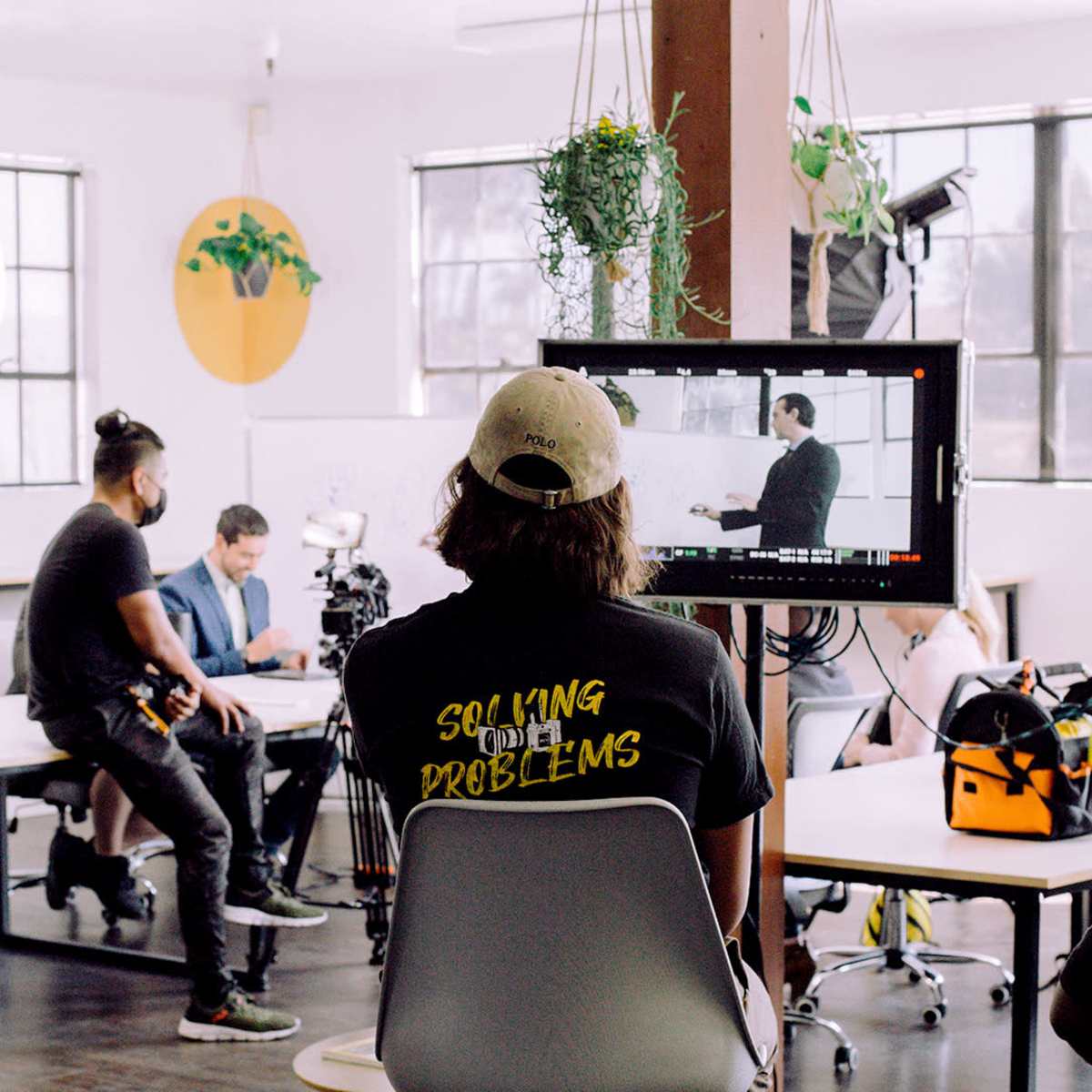 A film crew works in a bright studio setting, setting up equipment while a subject rehearses in front of a camera.
