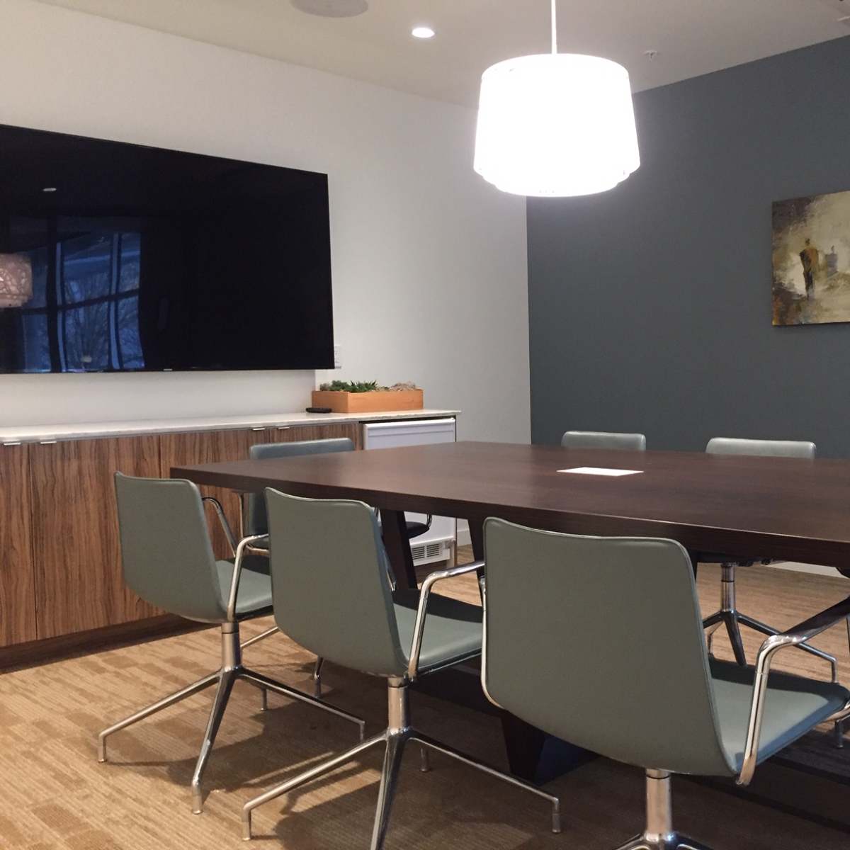 A modern conference room features a large wooden table surrounded by six light blue chairs, a wall-mounted TV, and an abstract painting.