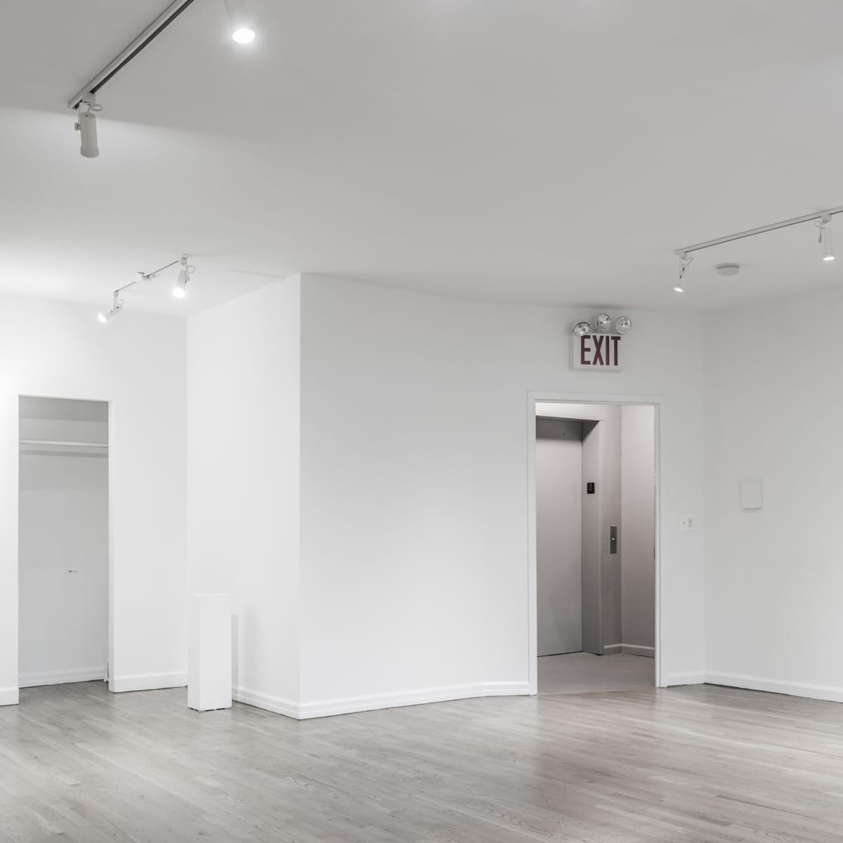 A minimalist, empty gallery space with white walls, wooden flooring, and spotlights illuminating the area.