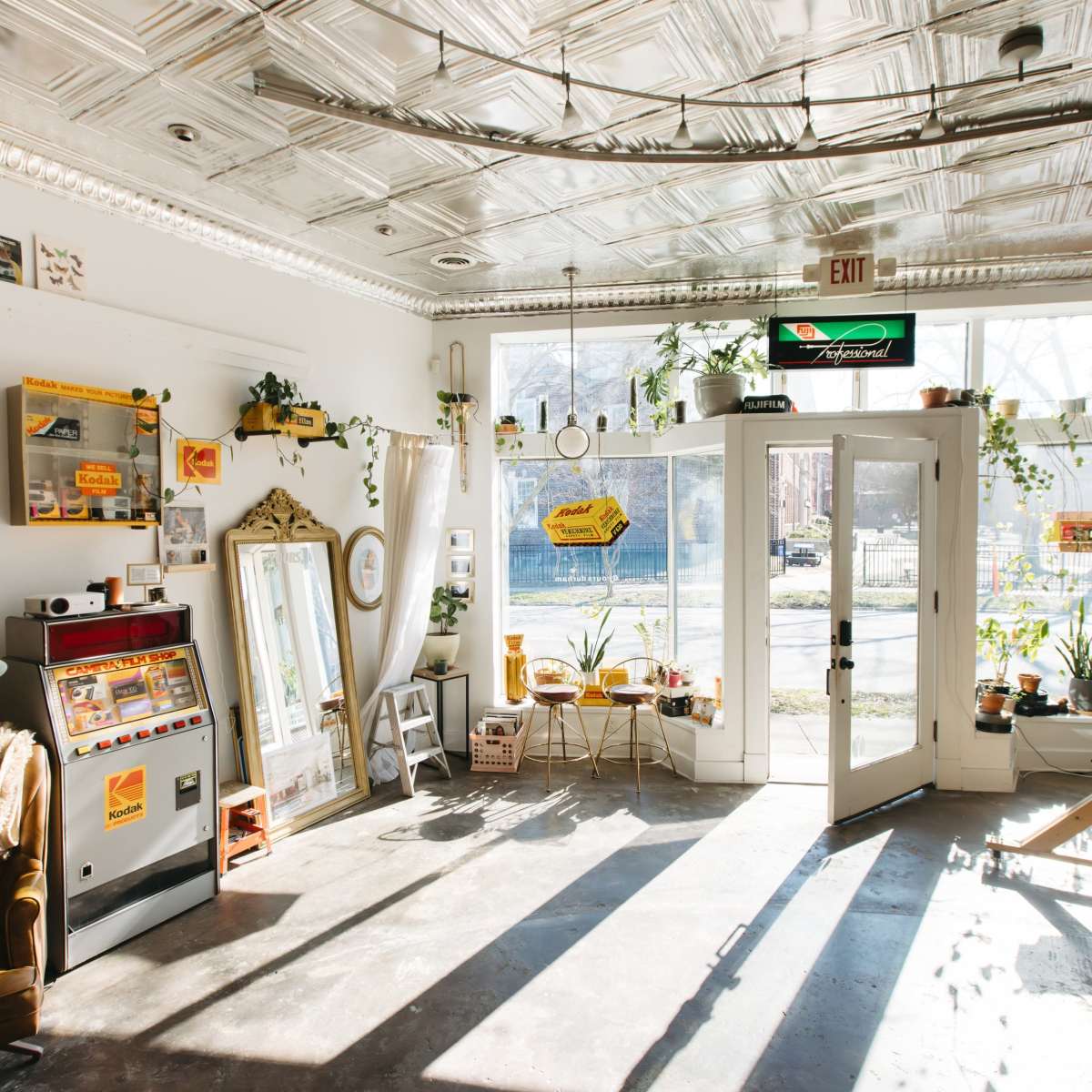 A bright, airy room with large windows, featuring a mix of vintage decor, plants, a seating area, and a vintage vending machine.
