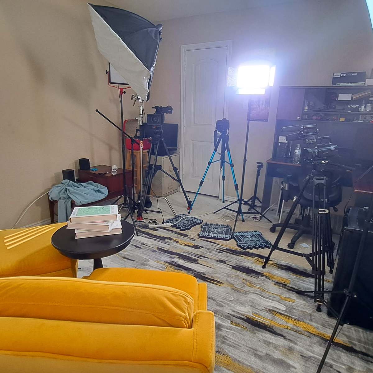 A brightly lit recording setup in a room featuring a yellow chair, a small table with scattered papers, and multiple cameras and tripods positioned around the space.
