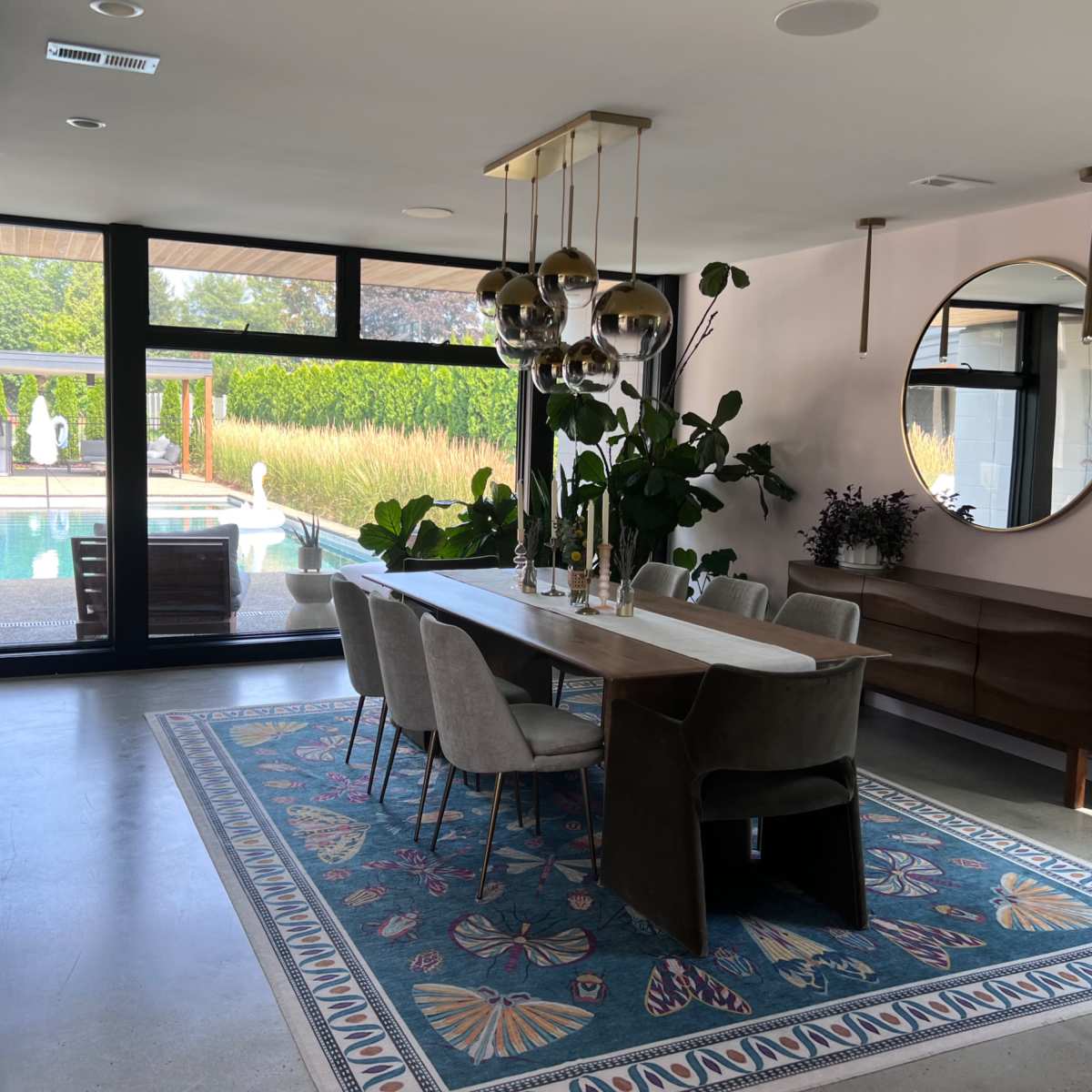 A dining area features a long table with six chairs, a large mirror, and plants, overlooking a pool through floor-to-ceiling windows.